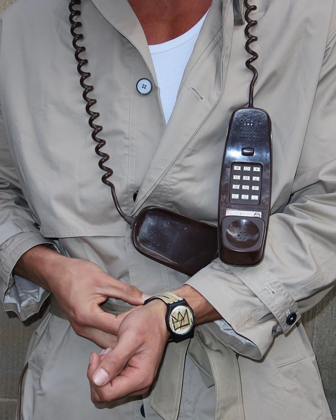 Adult man wearing a tan trench coat, white undershirt, and a watch with a crown design. A vintage phone hangs across his chest.