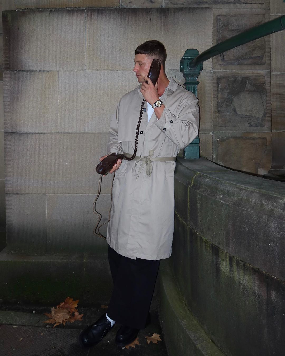 A man in a trench coat stands outdoors using a vintage telephone with a curly cord near a stone wall and greenery.