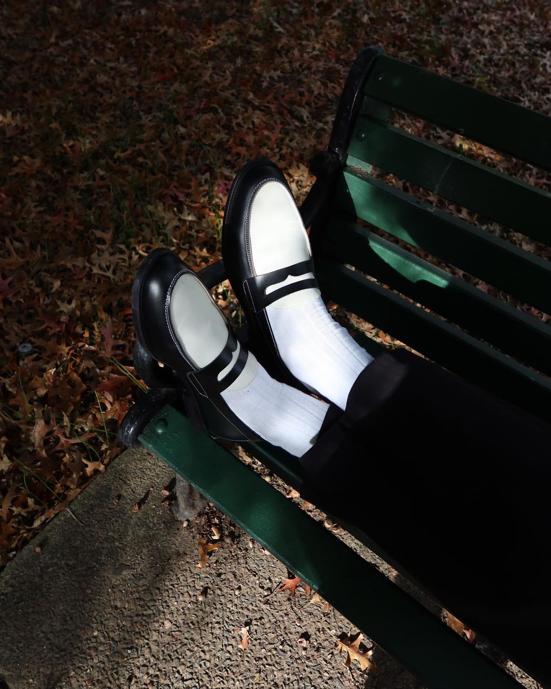 A pair of black and white spectator-style shoes worn with white socks on a green park bench outdoors in dappled sunlight.