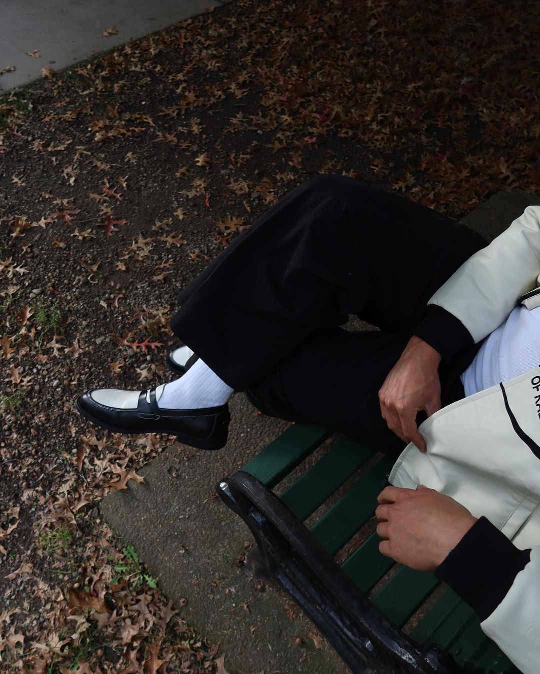 Person resting on a green park bench in black pants, white socks, and black and white loafers, amidst fallen leaves during the day.