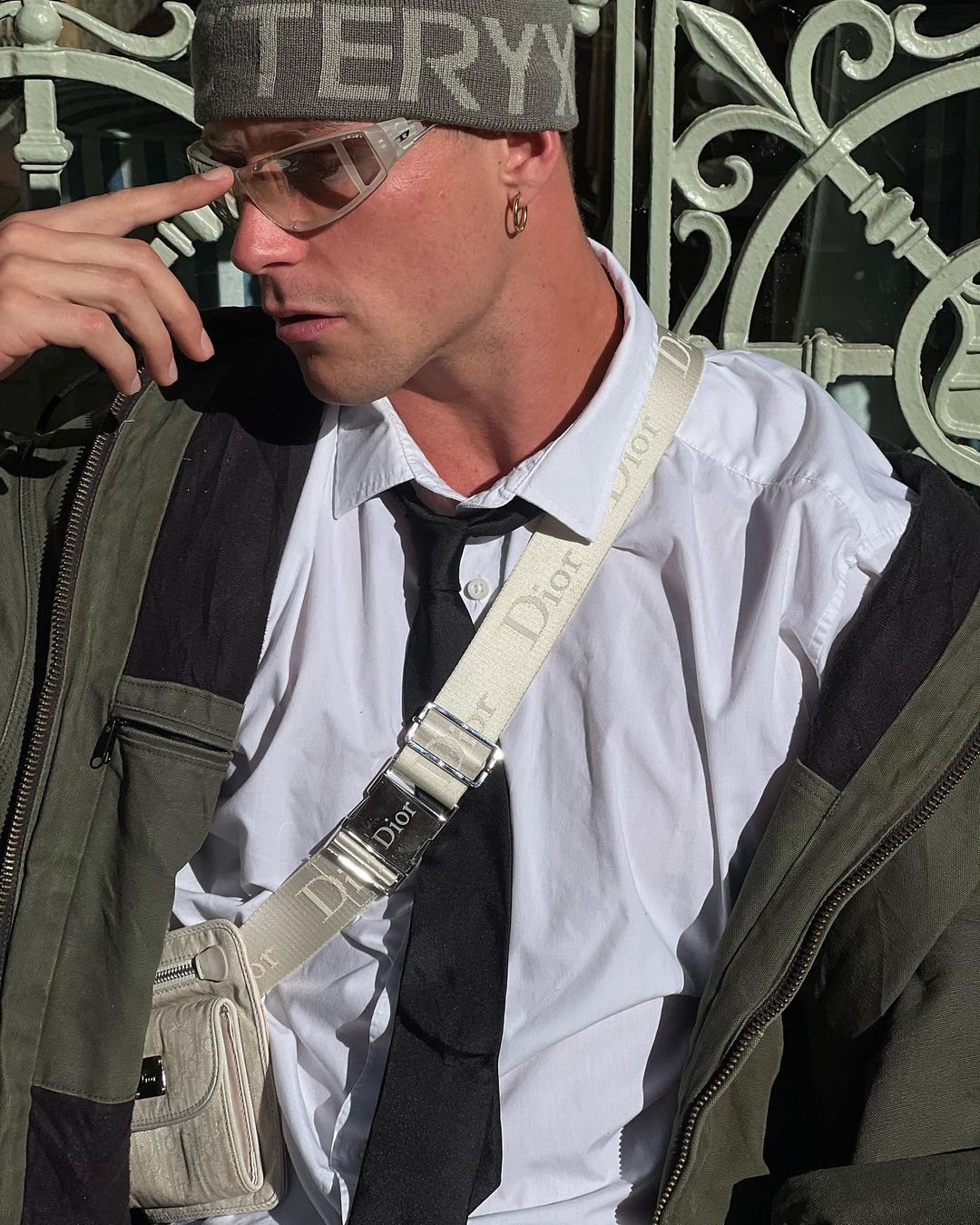 Stylish man in beanie, glasses, and Dior bag standing outdoors near a wrought iron fence, with a white dress shirt and tie.