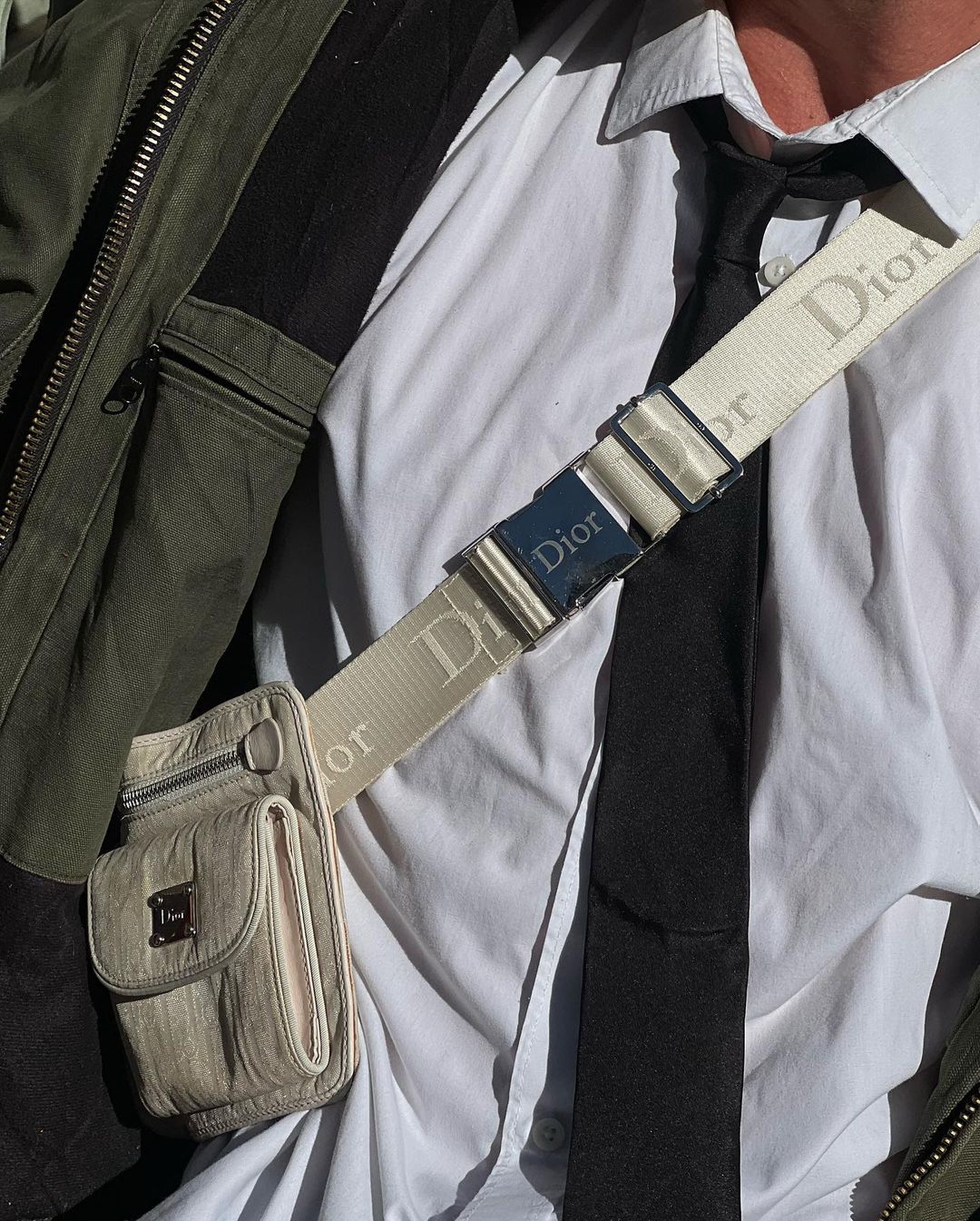 Close up of a person wearing a white button-down shirt, black tie, and a beige crossbody bag with a Dior branded strap and buckle.