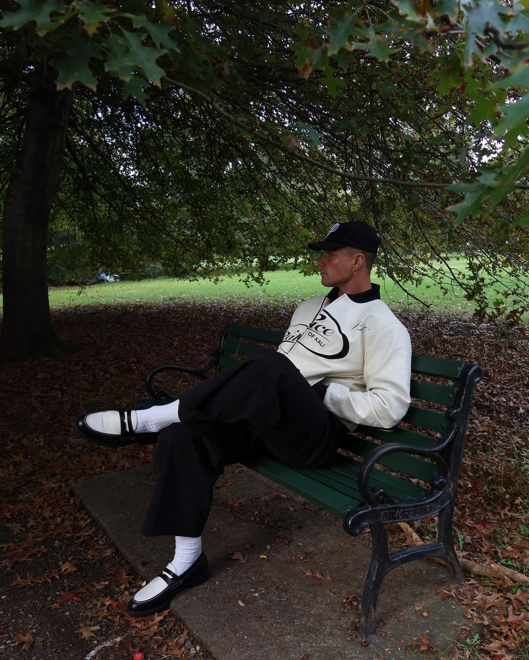 Man sits on a park bench beneath a tree, wearing a vintage-style sweater, black pants, white socks, and black and white loafers.