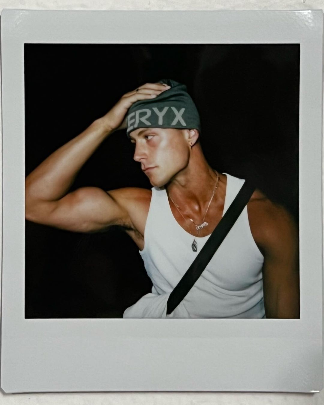 A muscular adult male poses wearing a tank top and beanie against a dark backdrop. He has a necklace and an over-the-shoulder bag.