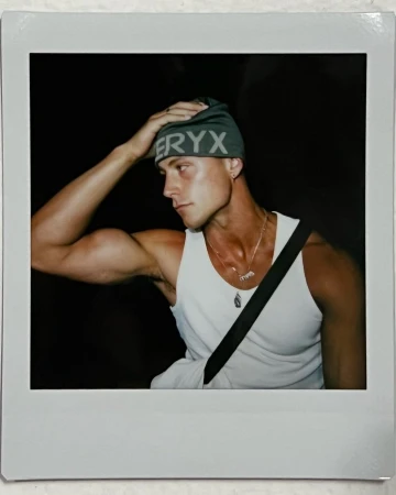 Polaroid photograph of a man in a white tank top and green beanie adjusting his hat against a dark background