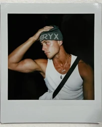 Polaroid photograph of a man in a white tank top and green beanie adjusting his hat against a dark background