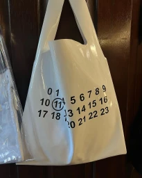 Glossy white plastic tote bag featuring black numeric print hanging against a dark wood panel door in Sydney by Will Stokoe.