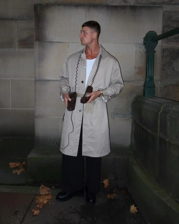 Man wearing a beige trench coat over a white shirt and black trousers holding two vintage telephone handsets