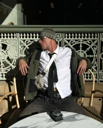 Man seated at a café wearing an olive green beanie, white shirt, black tie, and dark jacket with a crossbody bag.