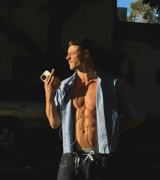 Man wearing unbuttoned striped shirt holding smartphone outdoors in harsh sunlight by Will Stokoe
