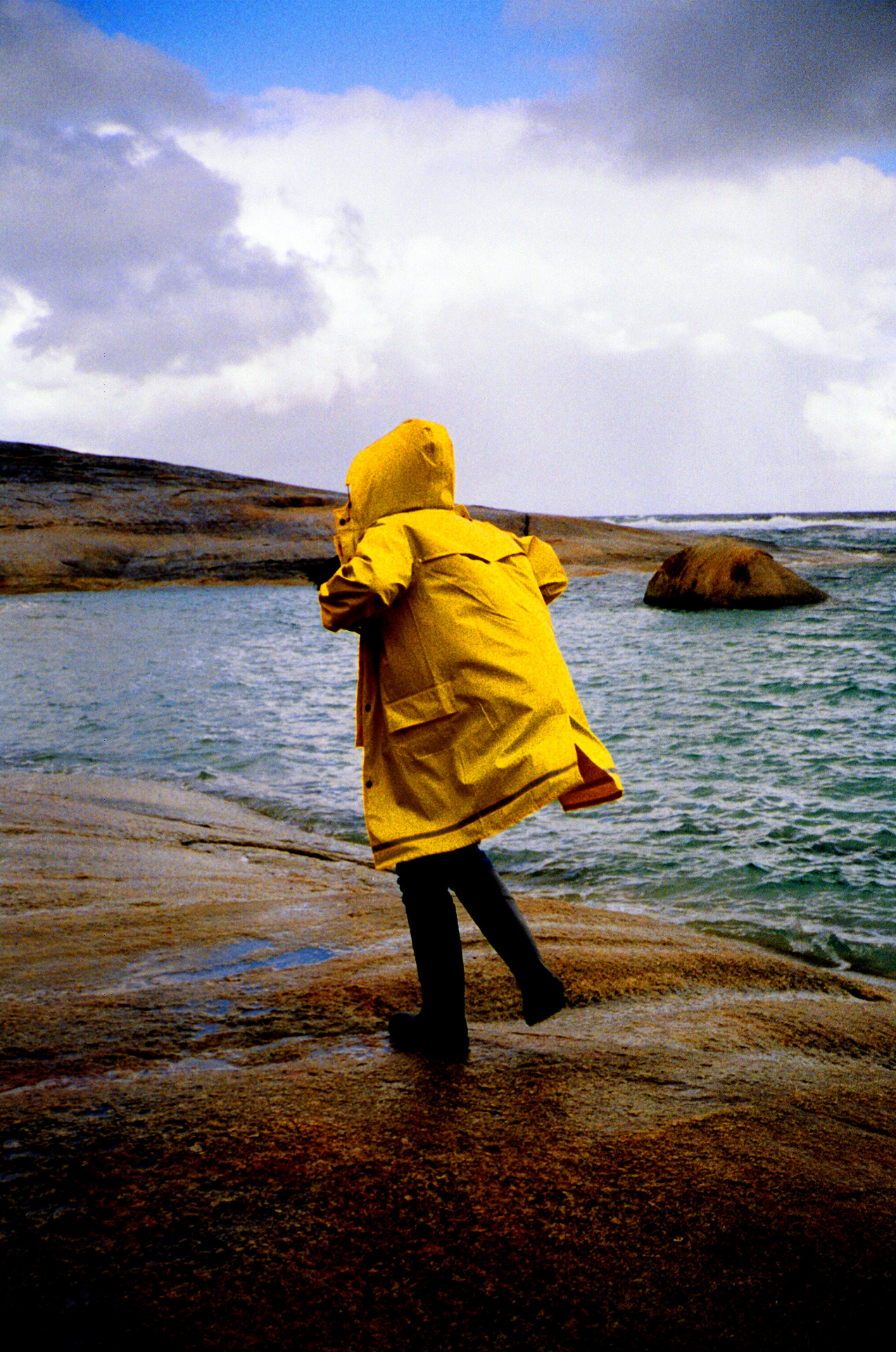 Person in yellow raincoat stands on rocky shore looking out at ocean under cloudy sky. Rainwear offers protection during outdoor weather.