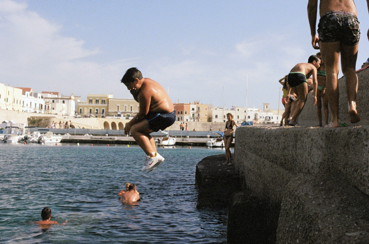 Boy jumping into ocean water near a concrete pier with other people; background shows light-colored buildings under blue sky, daytime.