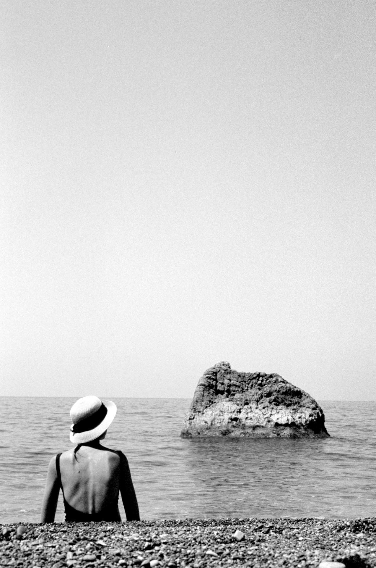 Back view of an adult woman sitting on a pebbled beach, wearing a hat, looking out at a rocky islet in the water on a clear day.