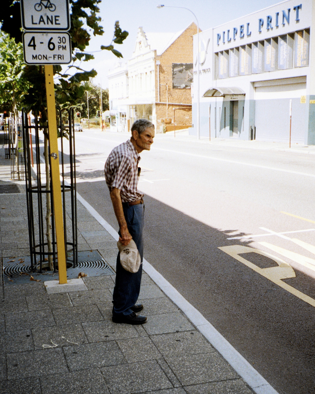An elderly man in a plaid shirt stands on a sidewalk next to a street sign and buildings on a sunny day with shadows on the asphalt.