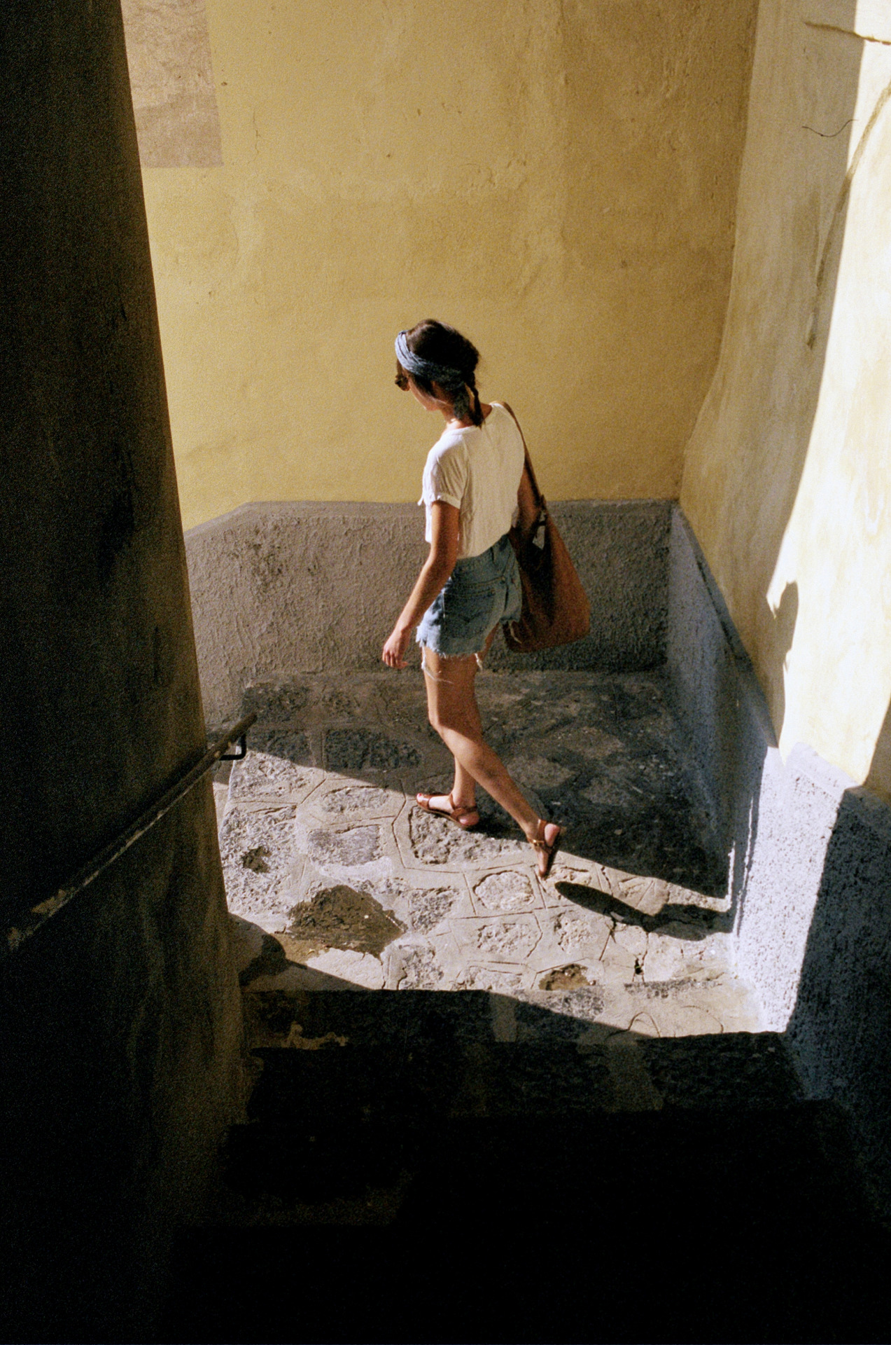 Woman in shorts and t-shirt descends stone stairs outdoors in sunlight, carrying a tote bag with a pillar visible to the left.