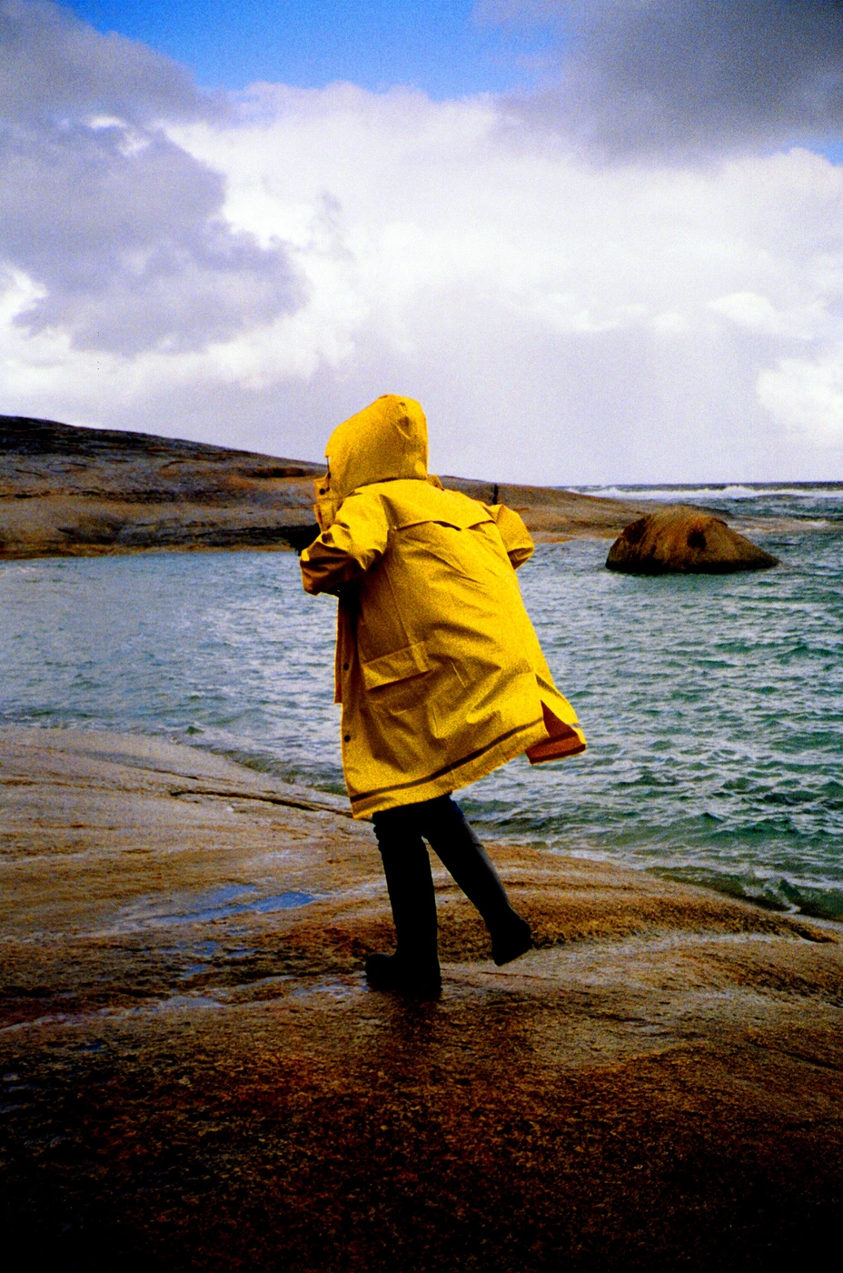 Person in a bright yellow hooded raincoat walking along a rocky coastal shore near the water under an overcast sky.
