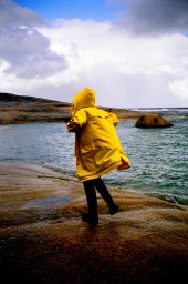 Person in a bright yellow hooded raincoat walking along a rocky coastal shore near the water under an overcast sky.
