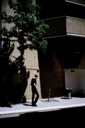 Man walking on a sidewalk past a brick building and a tree casting shadows in Sydney, captured by Will Braden.