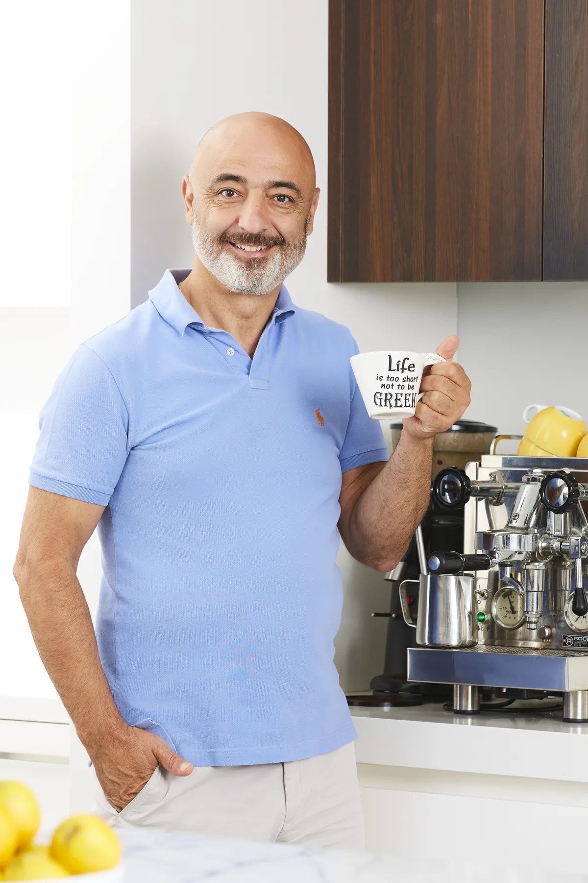Smiling mature man in polo shirt holds coffee mug with text, posing in a bright indoor kitchen with an espresso machine visible.