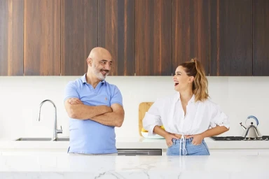 Man and woman smiling in a modern kitchen with dark wood upper cabinets and white countertops.
