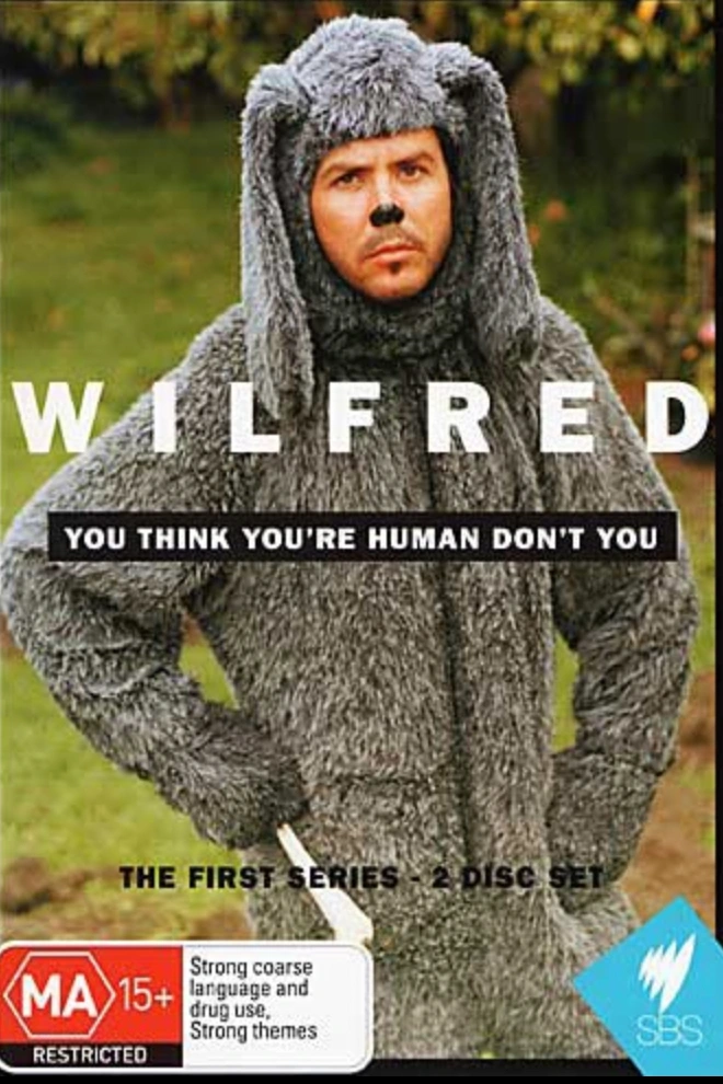 Man wearing a shaggy grey dog suit standing outdoors with text reading WILFRED above him
