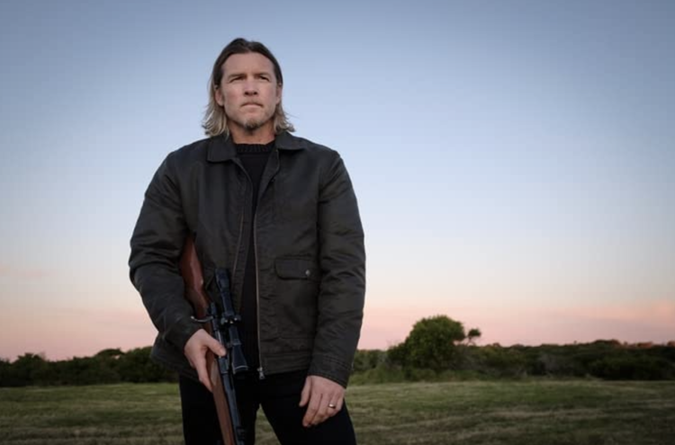 Adult male poses outdoors at dusk holding a rifle, wearing a dark jacket, in a field with trees and a twilight sky.