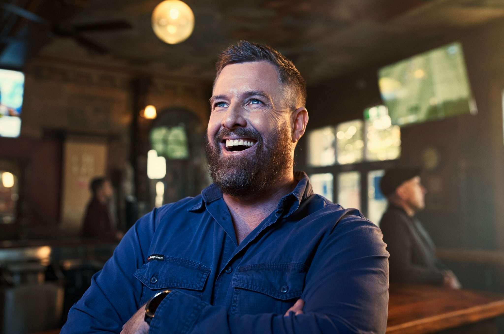 Adult man smiling with arms crossed in a casual blue button shirt, inside a bar setting with bar and patrons in soft focus.