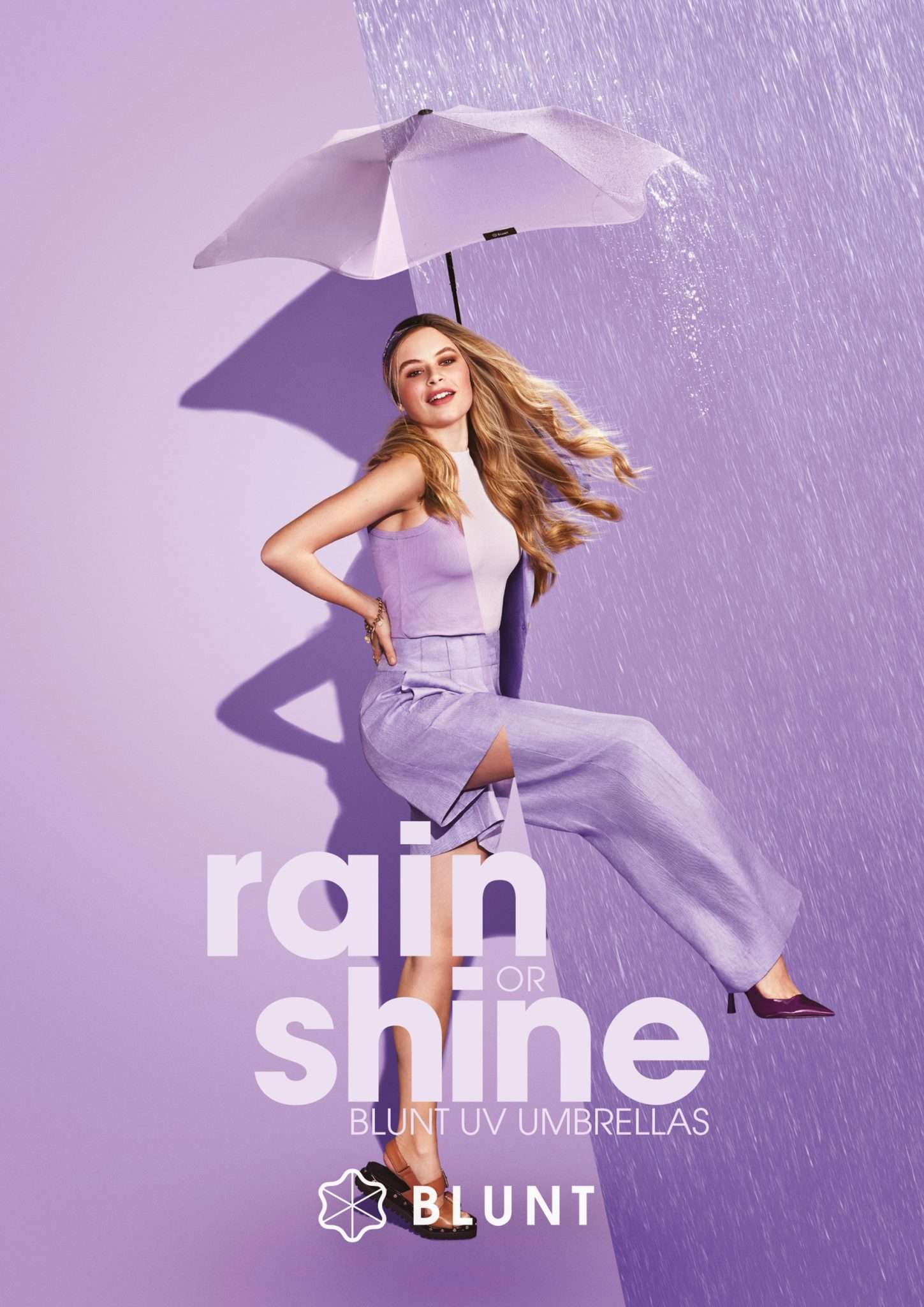 A young woman poses with an umbrella, standing against a dual-tone lilac backdrop with simulated rain, promoting Blunt UV umbrellas.