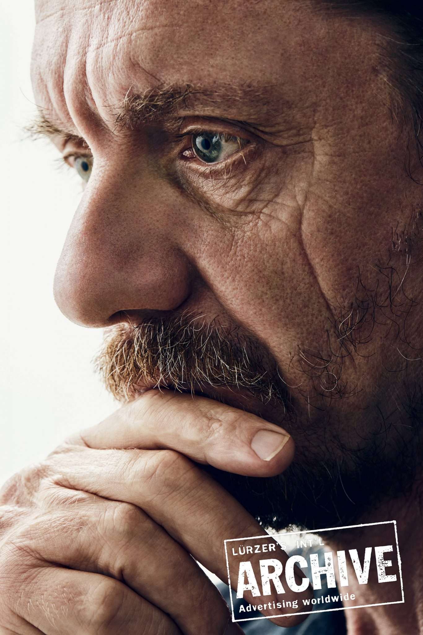 Close-up portrait of a mature man with a beard, his hand thoughtfully touching his face, against a bright background and Lurzer's Archive logo.