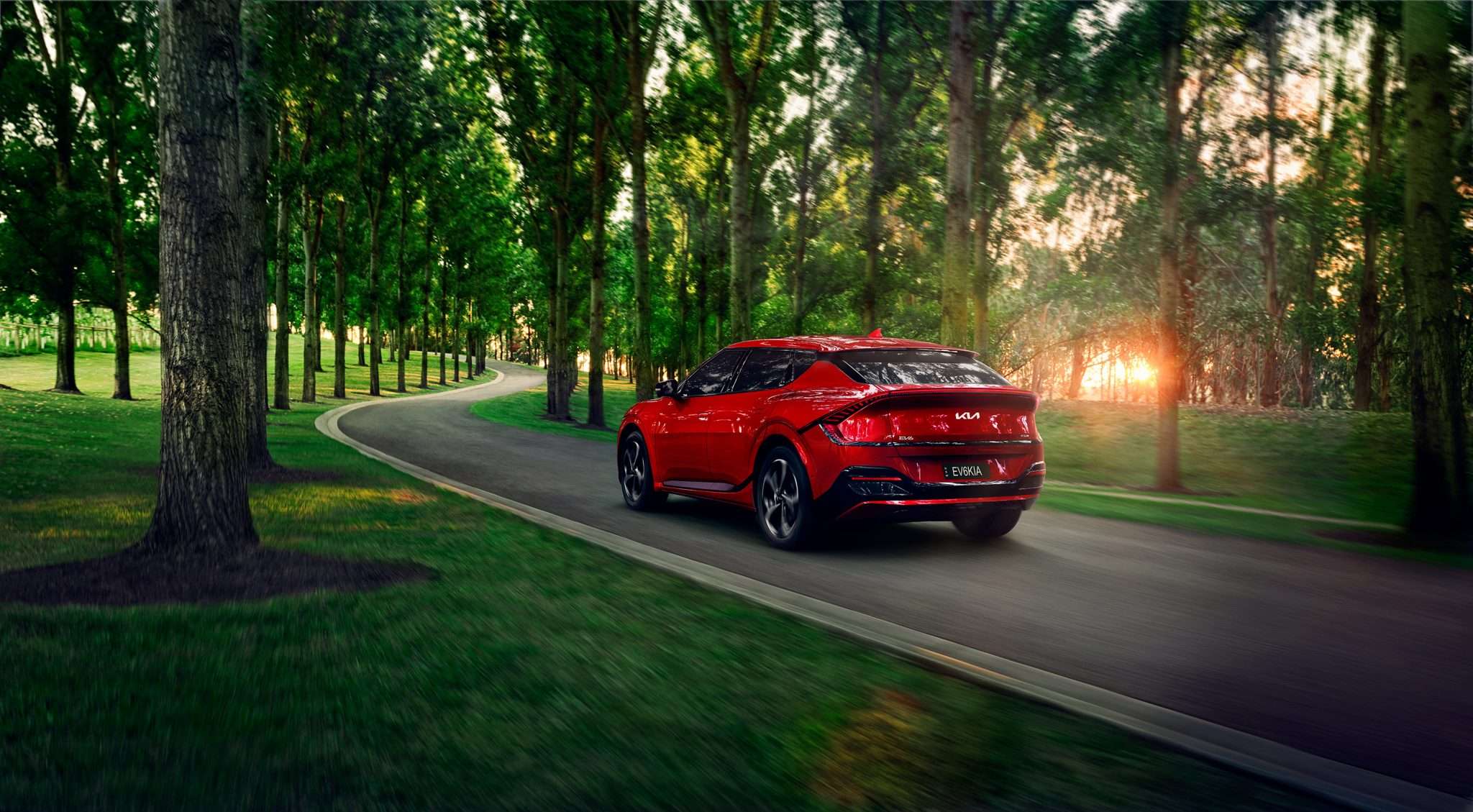 Rear view of a red Kia EV6 driving on an asphalt road lined with tall trees and green grass, showcasing automotive design and scenic environment.