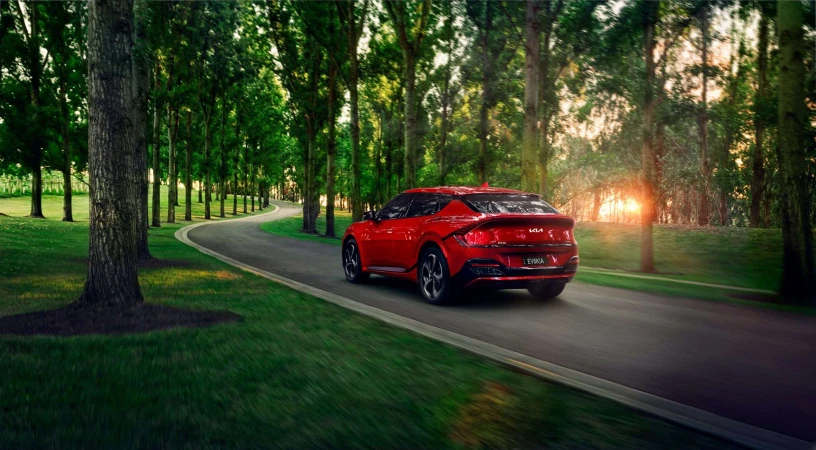 Rear view of a red Kia EV6 driving on an asphalt road lined with tall trees and green grass, showcasing automotive design and scenic environment.