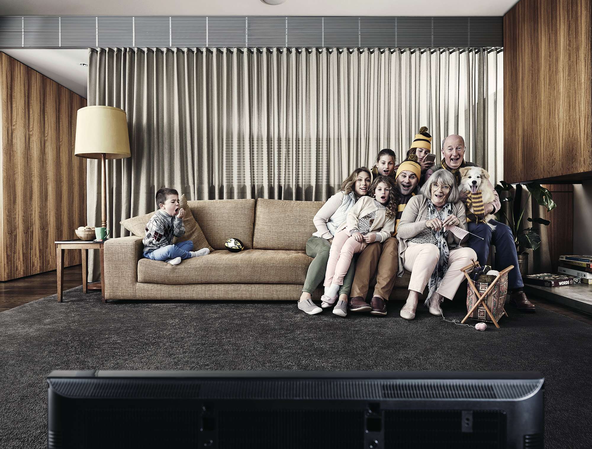 Family of eight, including two children, and a dog, are sitting on a couch in living room watching television, lit by a floor lamp.