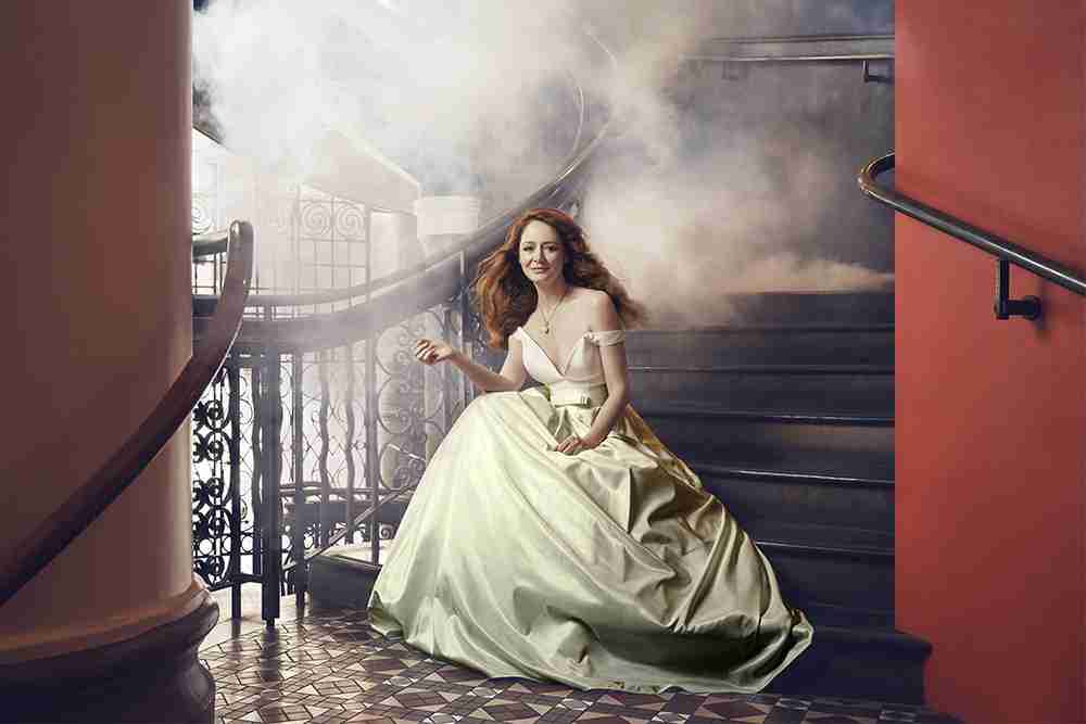 Elegant woman in a formal gown sitting on stairs in an indoor setting, surrounded by decorative ironwork and soft lighting.