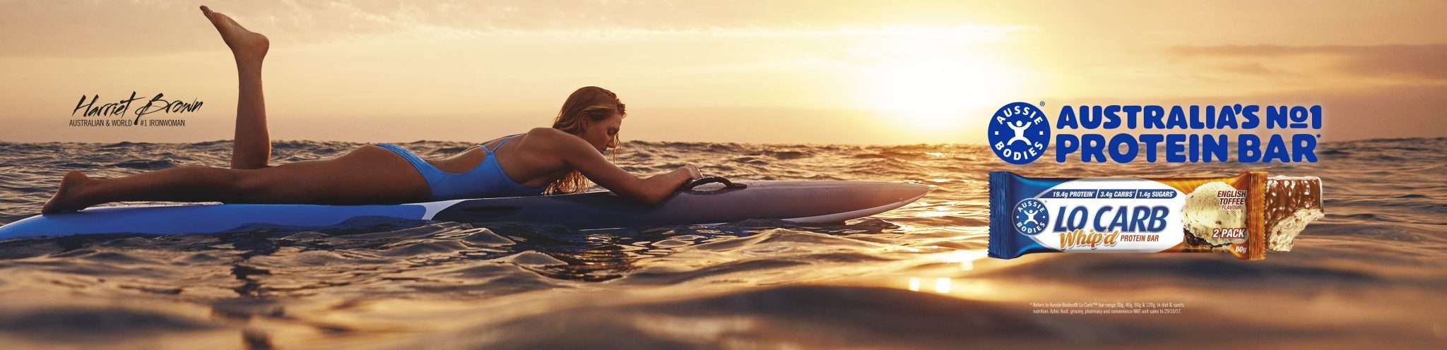 A woman in a blue swimsuit is laying on a surfboard in the ocean with Aussie Bodies protein bars visible against a bright sunset sky.