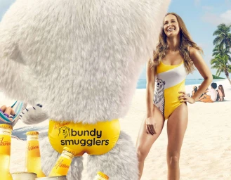 Giant white fuzzy bear wearing yellow swim briefs next to a smiling woman in a yellow and white swimsuit on a sandy beach