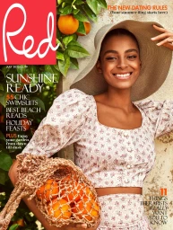 Smiling woman in a wide-brimmed sunhat holding a net bag filled with oranges against an old stone wall with citrus branches.