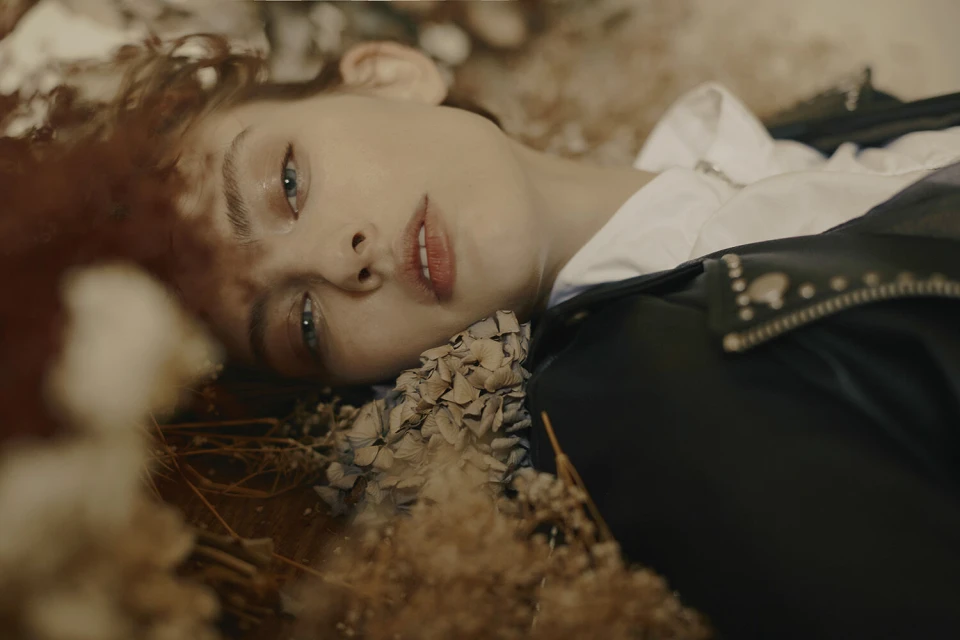 Portrait of a woman lying in dried flowers and greenery wearing a black leather jacket and white shirt in an indoor setting.
