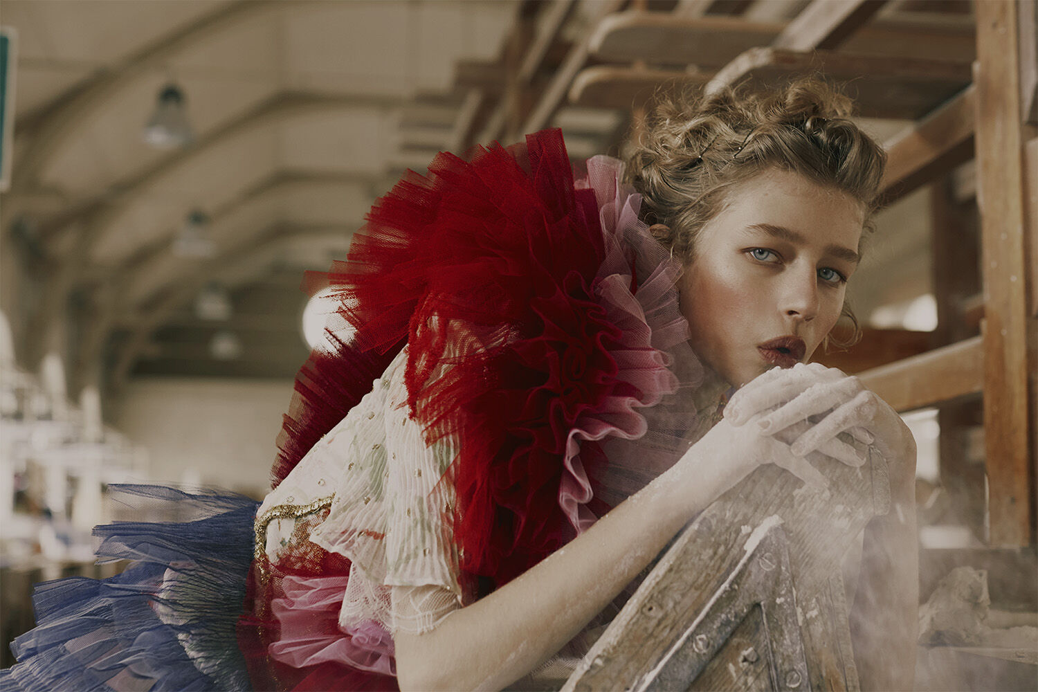 Young woman in colorful, ruffled couture fashion poses in a rustic indoor location with a pensive expression and powdered hands.