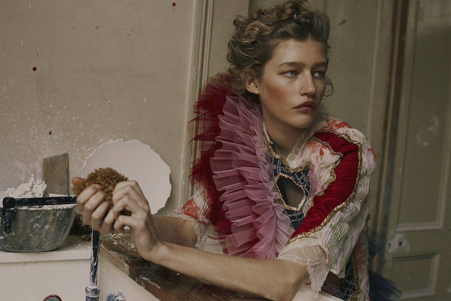 Portrait of a young woman wearing an elaborate ruffled collar and makeup, holding a sponge near a sink in a worn indoor setting.