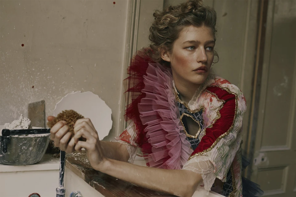 Portrait of a young woman wearing an elaborate ruffled collar and makeup, holding a sponge near a sink in a worn indoor setting.