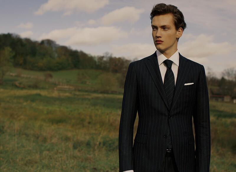 Adult male in pinstripe suit and tie stands in a field of green grass with a hill and blue sky with clouds in the background.