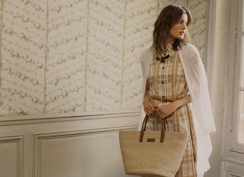 A fair-skinned adult woman with brown hair holds a woven tote bag while wearing a plaid midi dress, a white jacket, and a brown belt indoors.