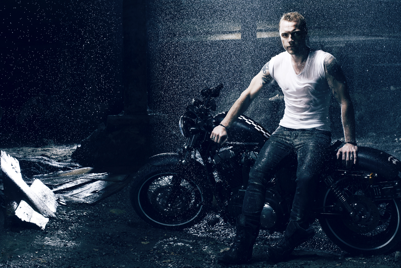 Man in white T-shirt poses on black motorcycle in the rain. Dark, moody atmosphere with visible water droplets and sleek design.