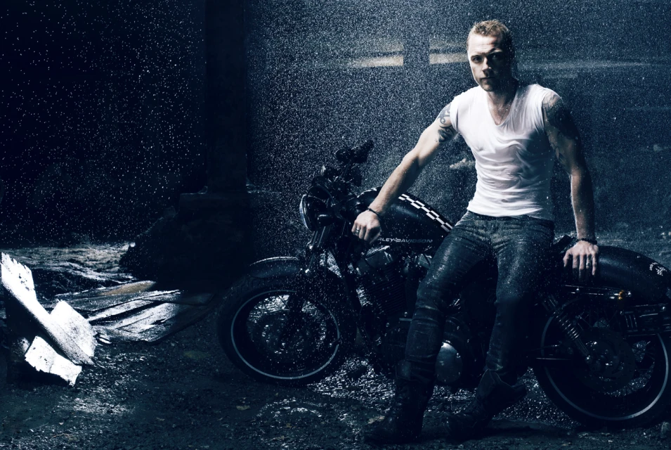 Man in white T-shirt poses on black motorcycle in the rain.  Dark, moody atmosphere with visible water droplets and sleek design.