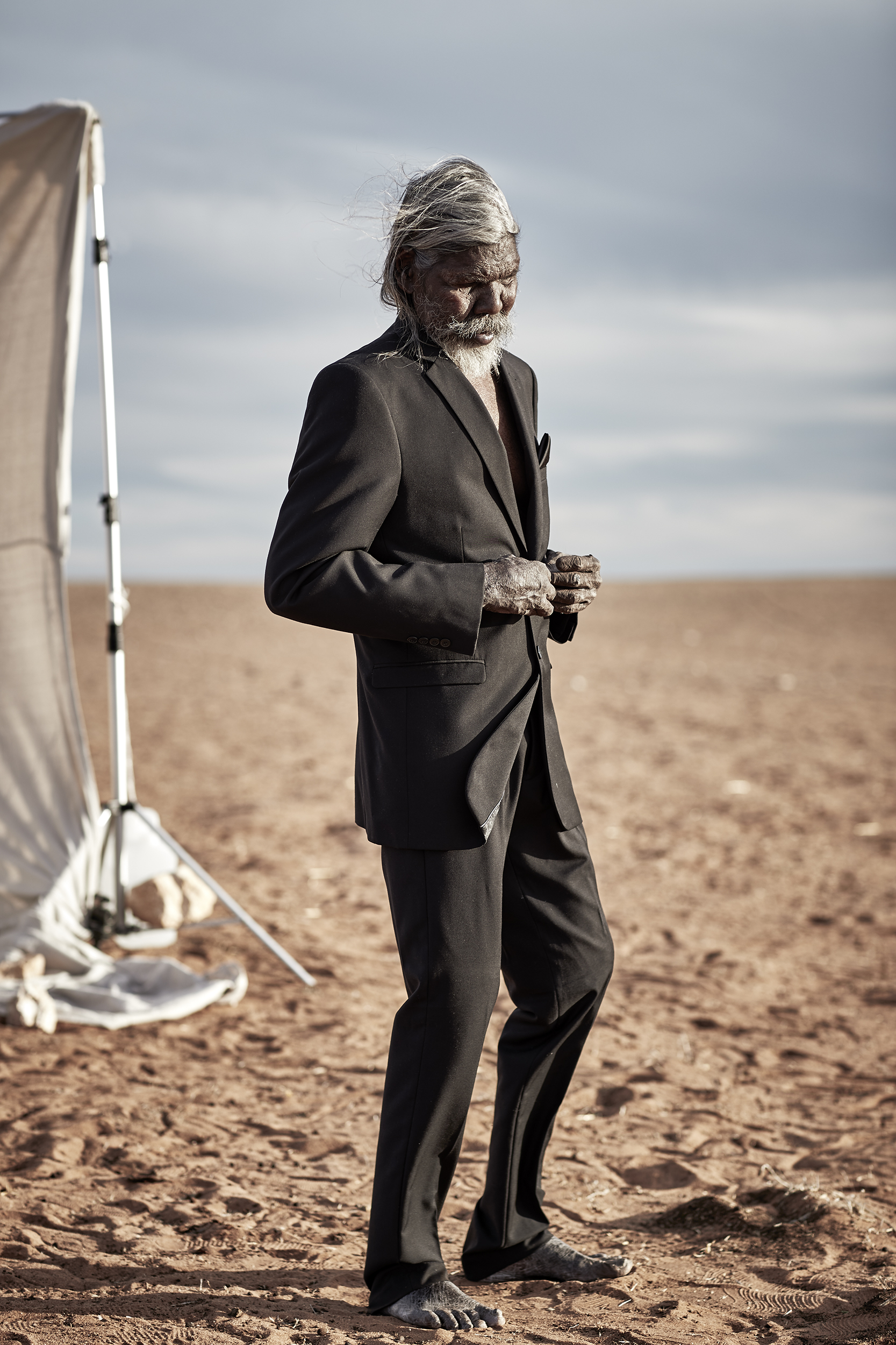 Elderly Indigenous man in a suit stands barefoot in a desert landscape. Blue sky and horizon visible behind him. Professional photo.
