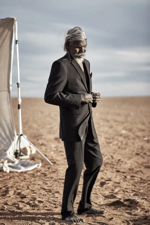 Elderly Indigenous man in a suit stands barefoot in a desert landscape. Blue sky and horizon visible behind him. Professional photo.
