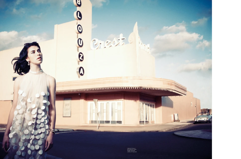 A woman in a dress stands in front of the Crest Theater under a bright blue sky with a classic car parked in the background.