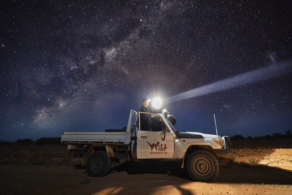 Person with headlamp next to 4x4 vehicle under a star-filled night sky, illuminated by a bright beam of light, creating a dramatic scene.