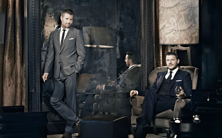 Man in grey suit standing beside another man seated in an armchair holding a glass in a dimly lit interior