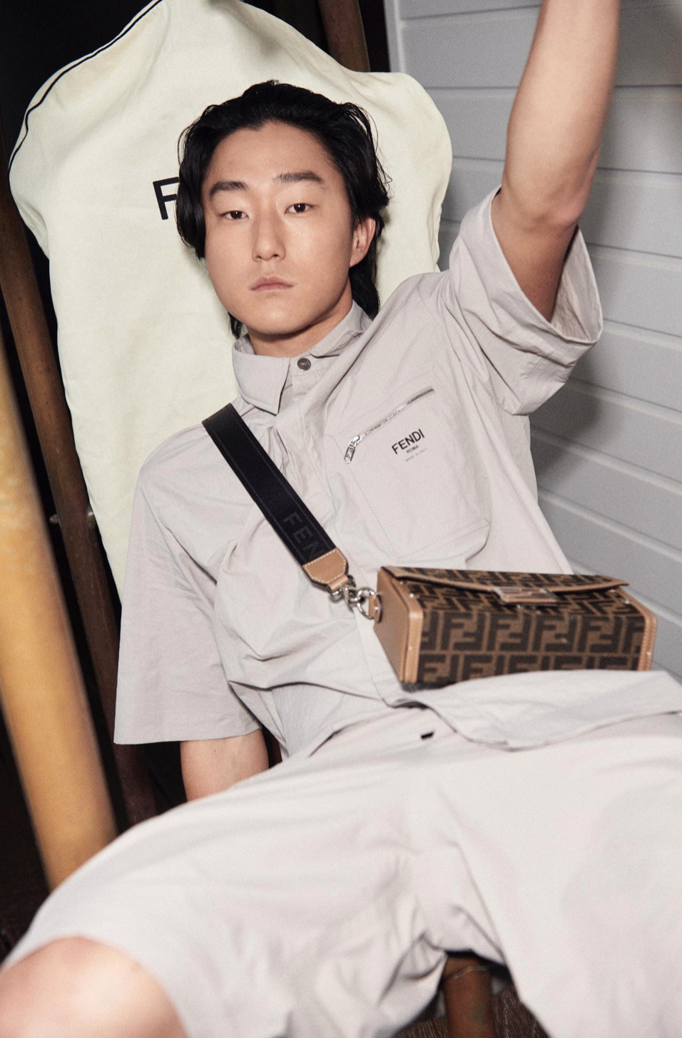 Young man in casual shirt and shorts sits, posing with a patterned brown Fendi handbag and arm raised indoors with natural lighting.